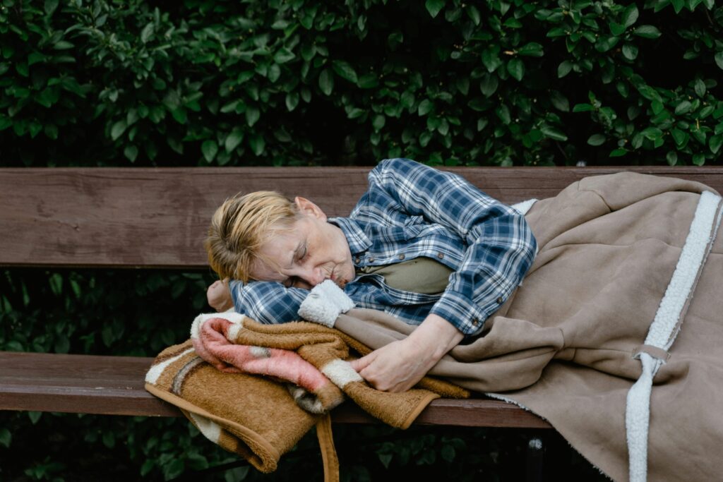 An elderly woman wearing a plaid shirt sleeps on a park bench covered with a blanket.
