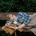 An elderly woman wearing a plaid shirt sleeps on a park bench covered with a blanket.