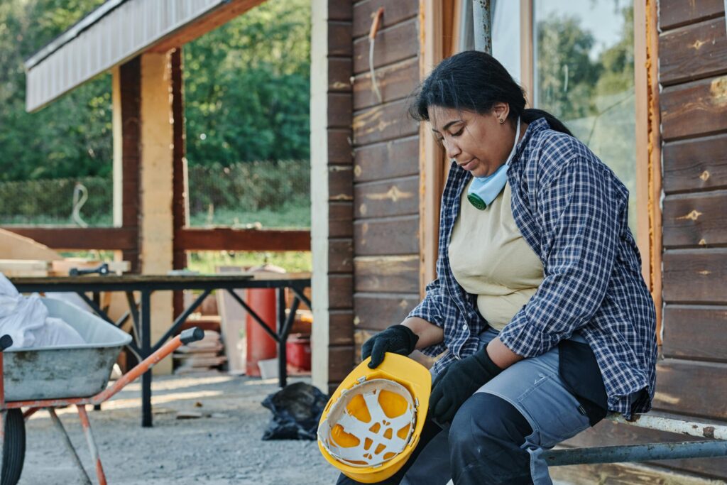A construction worker in a plaid shirt and hard hat takes a break outdoors, reflecting on the day.
