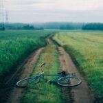 A vintage bicycle lies on an unpaved rural path amidst green fields under a cloudy sky.