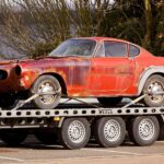 A worn-out, rusty vintage car secured on a flatbed trailer, outdoors under daylight.