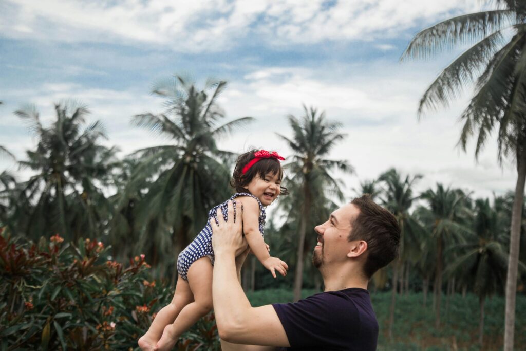 Smiling father lifting baby girl in tropical outdoors, surrounded by palm trees.