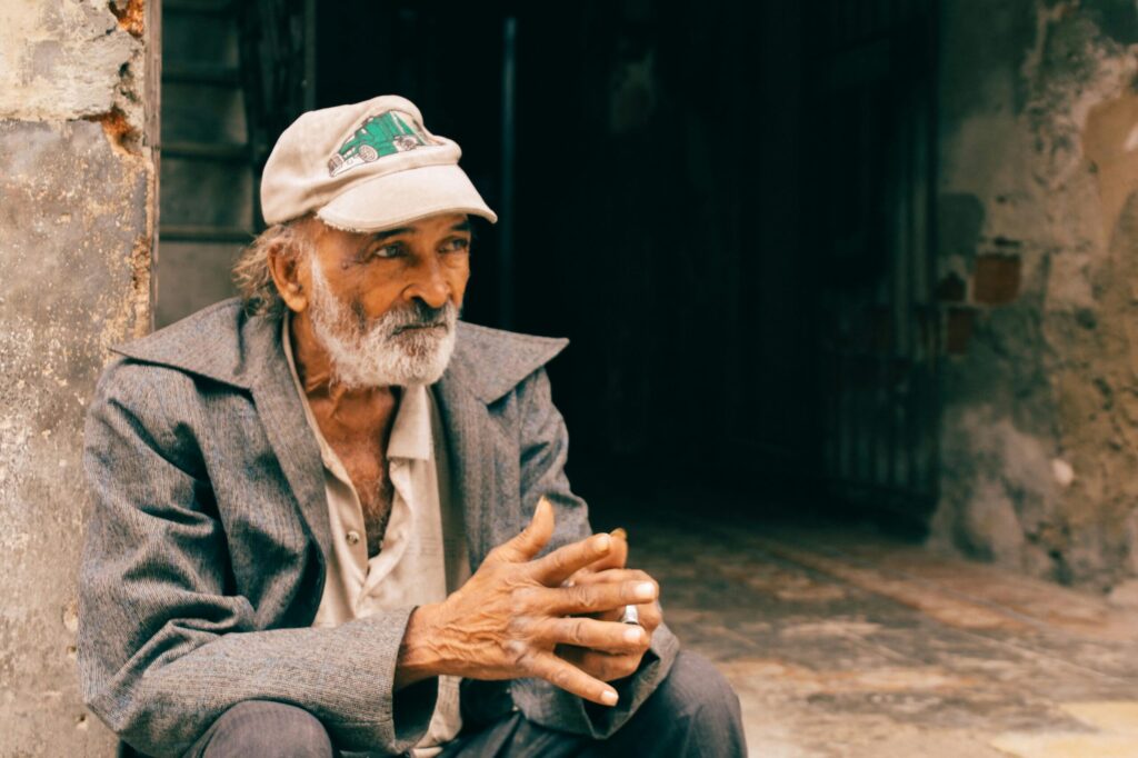 Elderly man in a cap sitting outdoors against a rustic wall, thoughtfully gazing.