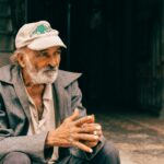 Elderly man in a cap sitting outdoors against a rustic wall, thoughtfully gazing.