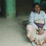 An elderly woman in traditional clothing sits outdoors, exuding cultural richness and simplicity.