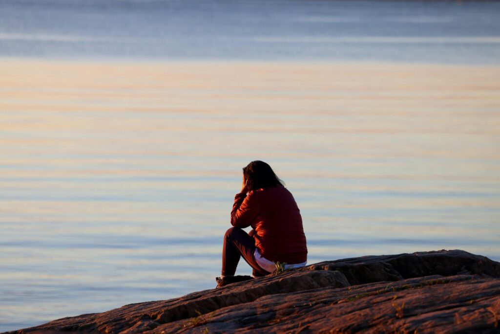 A woman in a red jacket sits on a rocky shore, contemplating the tranquil sea at sunset.