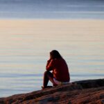 A woman in a red jacket sits on a rocky shore, contemplating the tranquil sea at sunset.