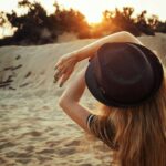 A woman wearing a fedora enjoys the serene sunrise at a sandy beach.