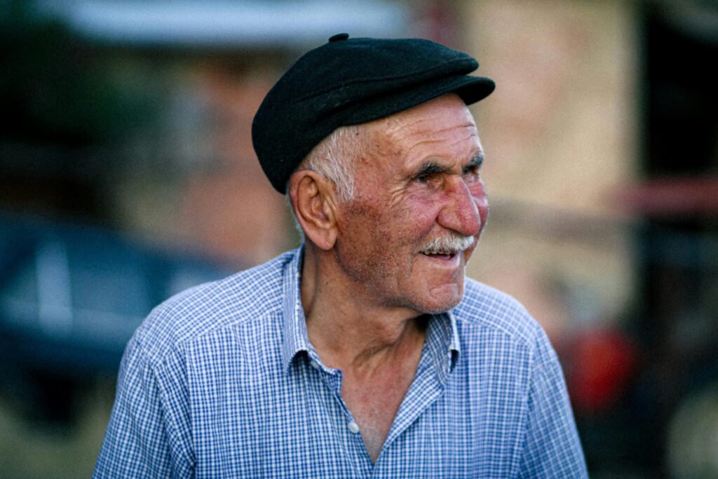 Portrait of a smiling elderly man wearing an ivy cap and checkered shirt outdoors.