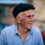 Portrait of a smiling elderly man wearing an ivy cap and checkered shirt outdoors.