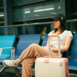 Young woman sitting in airport with luggage, anticipating travel. Casual travel attire.