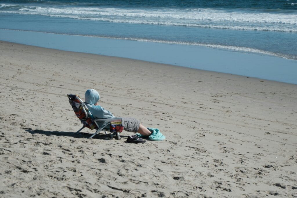 A person in a hoodie sits on a beach chair by the ocean, enjoying a sunny day.