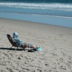 A person in a hoodie sits on a beach chair by the ocean, enjoying a sunny day.
