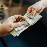 A close-up image of hands exchanging US dollar bills, symbolizing financial transaction or payment.