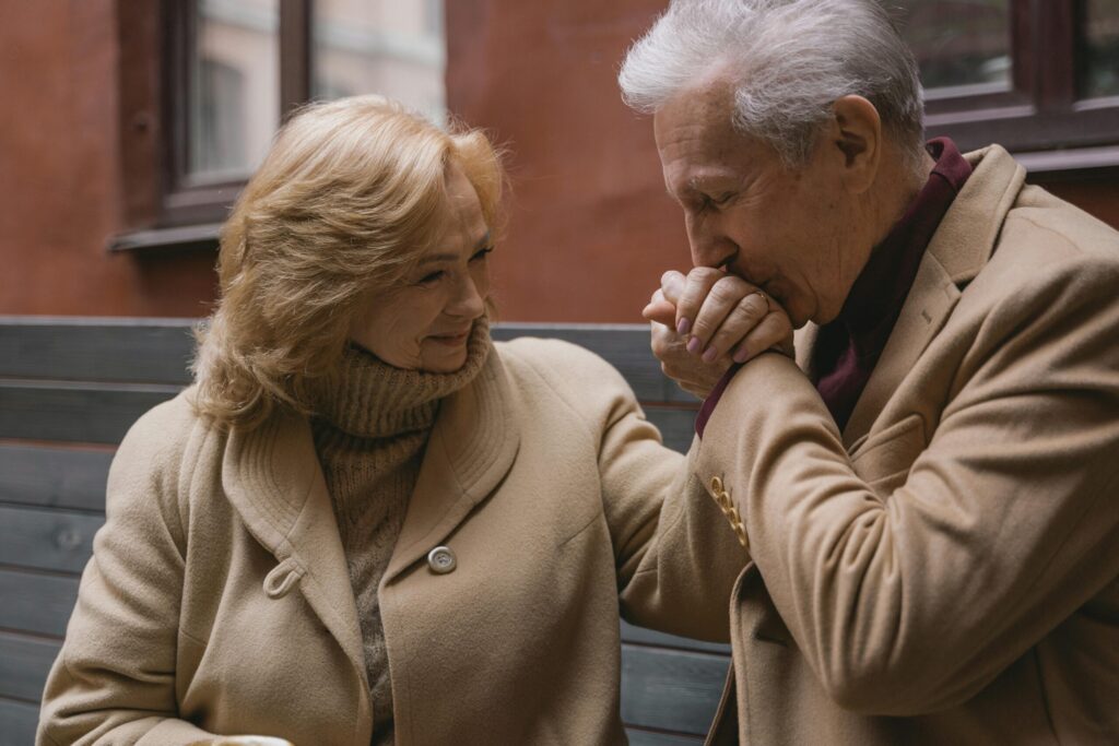 Elderly couple in beige coats sharing a loving moment, holding hands and smiling outdoors.
