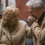 Elderly couple in beige coats sharing a loving moment, holding hands and smiling outdoors.