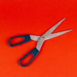 A pair of scissors with blue handles lies open on a bright orange background.