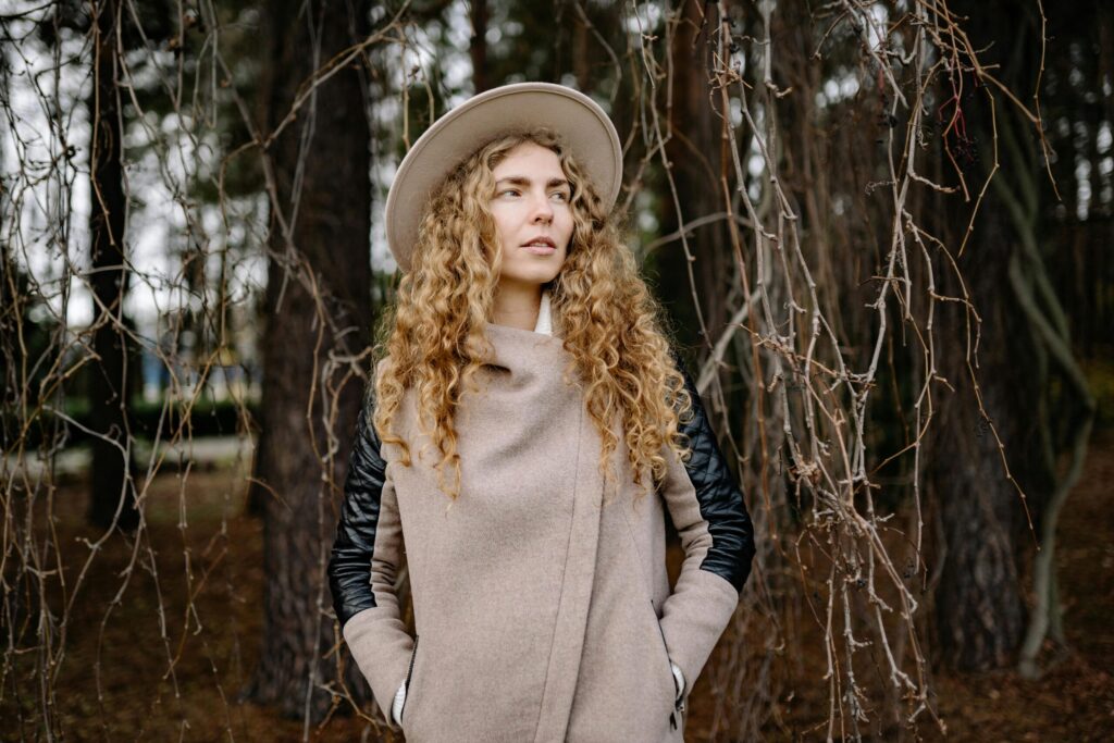 A woman with curly hair wearing a coat and hat, standing in an autumn forest setting with trees.