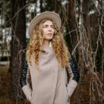 A woman with curly hair wearing a coat and hat, standing in an autumn forest setting with trees.