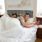 A couple relaxing in bed, with a woman using a smartphone in a cozy Portugal bedroom setting.