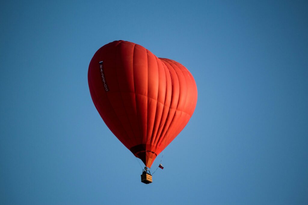 A striking red heart-shaped hot air balloon floats gracefully against a clear blue sky.