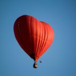 A striking red heart-shaped hot air balloon floats gracefully against a clear blue sky.