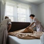 Two housekeepers making a bed in a sunlit bedroom, showcasing cleanliness and attention to detail.