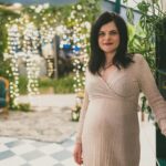 Pregnant woman in elegant dress standing indoors with soft lighting and greenery.