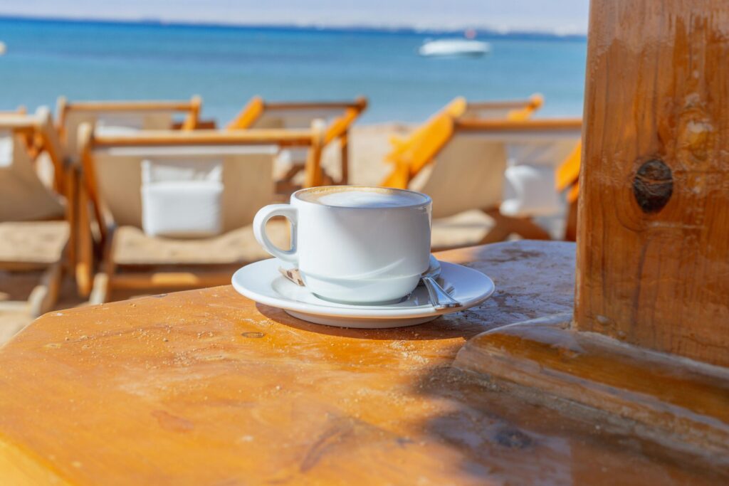 White cup of cappuccino on a wooden table with a blurry beach and sea background. Perfect for summer vibes.