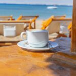 White cup of cappuccino on a wooden table with a blurry beach and sea background. Perfect for summer vibes.