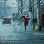 A busy urban street during heavy rain, with cars, pedestrians, and umbrellas.