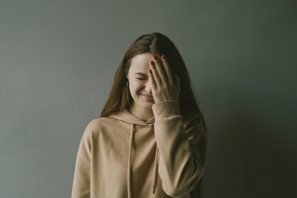 A young woman in a hoodie smiles with her hand covering part of her face against a neutral background.