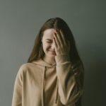 A young woman in a hoodie smiles with her hand covering part of her face against a neutral background.