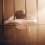 A tattooed man leans against prison bars, illuminated by dramatic sunbeams. Moody photography.
