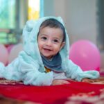 Happy baby in a hooded outfit crawling indoors surrounded by pink balloons.
