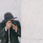 A person with a camera and gray hat capturing photos against a marble wall.