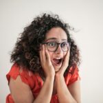A woman expresses surprise, her curly hair framing her eyeglass-wearing face, hands on cheeks.