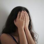 An adult woman with long brunette hair covers her face with her hands against a white wall, creating a minimalist portrait.