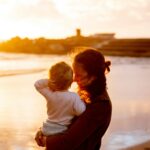 A loving mother embraces her child on a sunlit beach during sunset, celebrating family and togetherness.