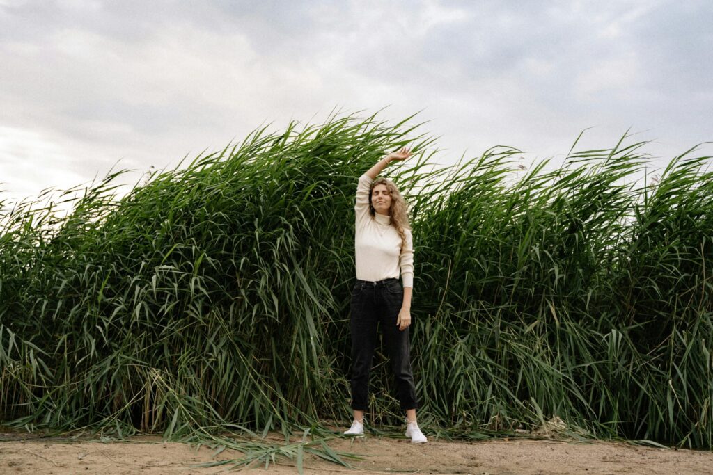 A woman with long hair poses with arm raised in front of tall grass and clouds.