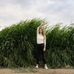 A woman with long hair poses with arm raised in front of tall grass and clouds.