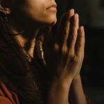 Close-up of a woman with folded hands praying, captured in a serene indoor setting with low lighting.