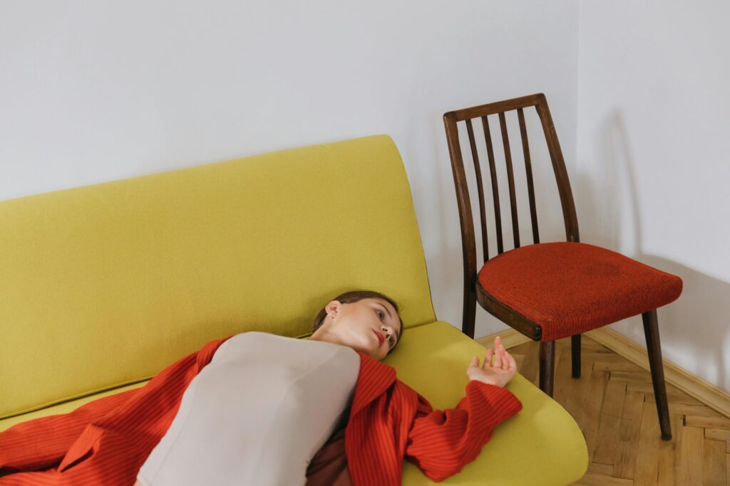 A woman in a red cardigan lying on a yellow couch beside a red chair indoors.