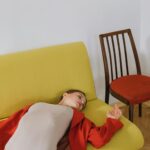 A woman in a red cardigan lying on a yellow couch beside a red chair indoors.