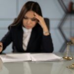 A fatigued female lawyer reviewing documents with a justice scale nearby, emphasizing stress in legal work.