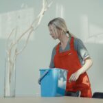 A woman in a red uniform cleans indoors with a blue bucket, ensuring a spotless environment.