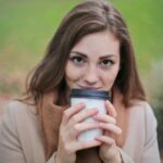 A young woman smiling while holding a coffee cup in a park during fall.