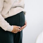 Close-up of a pregnant woman in black pants and beige top holding her belly.