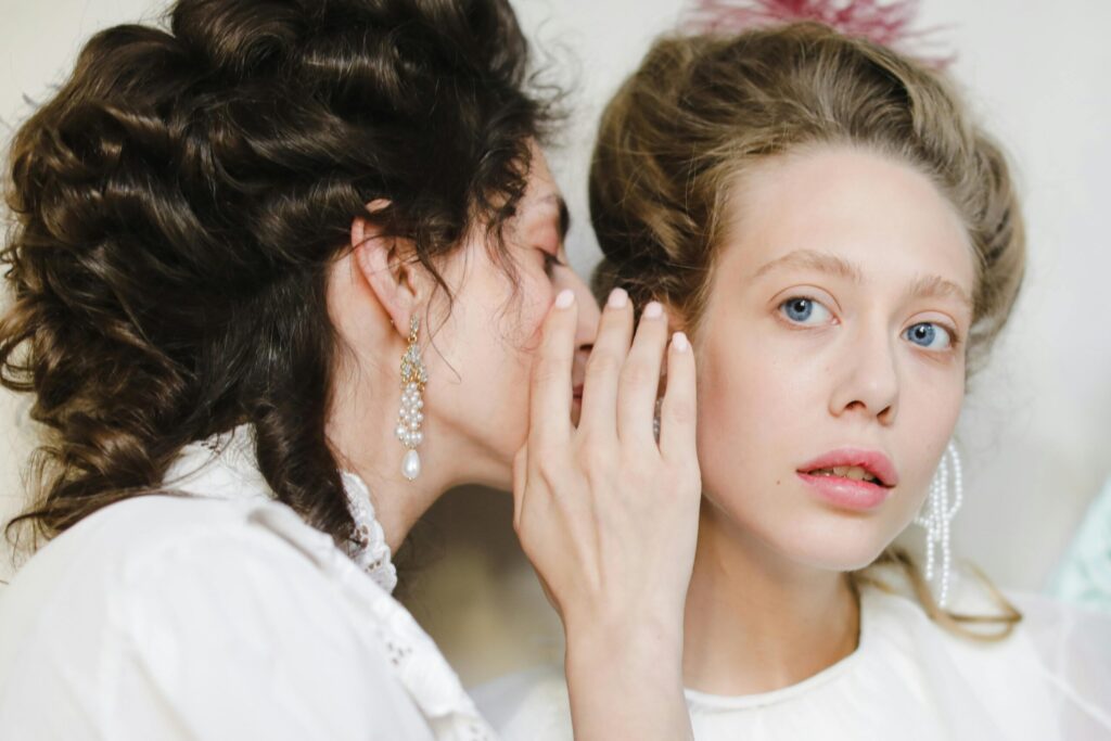Two women dressed in 19th century fashion share a secret, showcasing elegance and friendship.