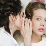 Two women dressed in 19th century fashion share a secret, showcasing elegance and friendship.